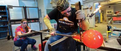 student clamping glass off of a pipe while another student blows to inflate the piece